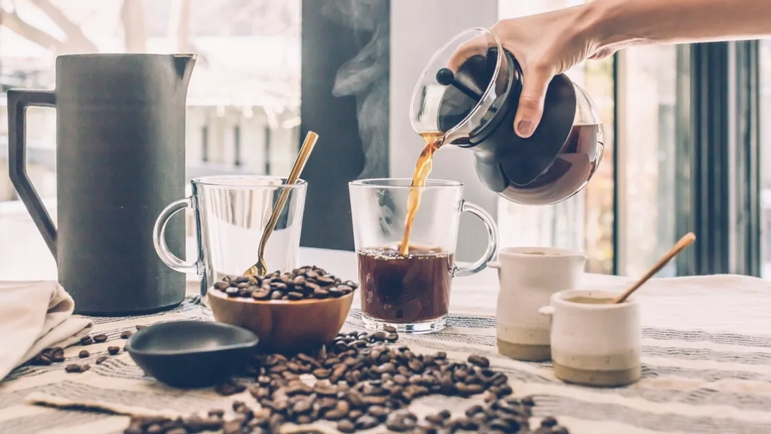Fresh coffee being poured from a glass carafe into a mug beside a bowl of roasted coffee beans. BMI Tracker - bmi-tracker.com