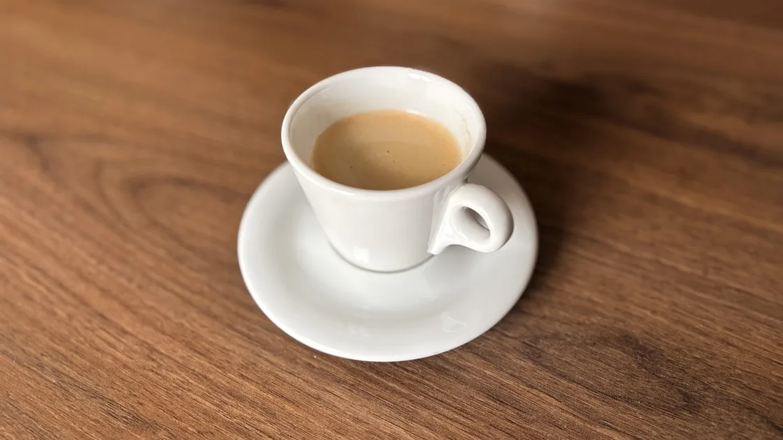 A small white ceramic espresso cup with crema on a saucer, set on a wooden table. BMI Tracker - bmi-tracker.com